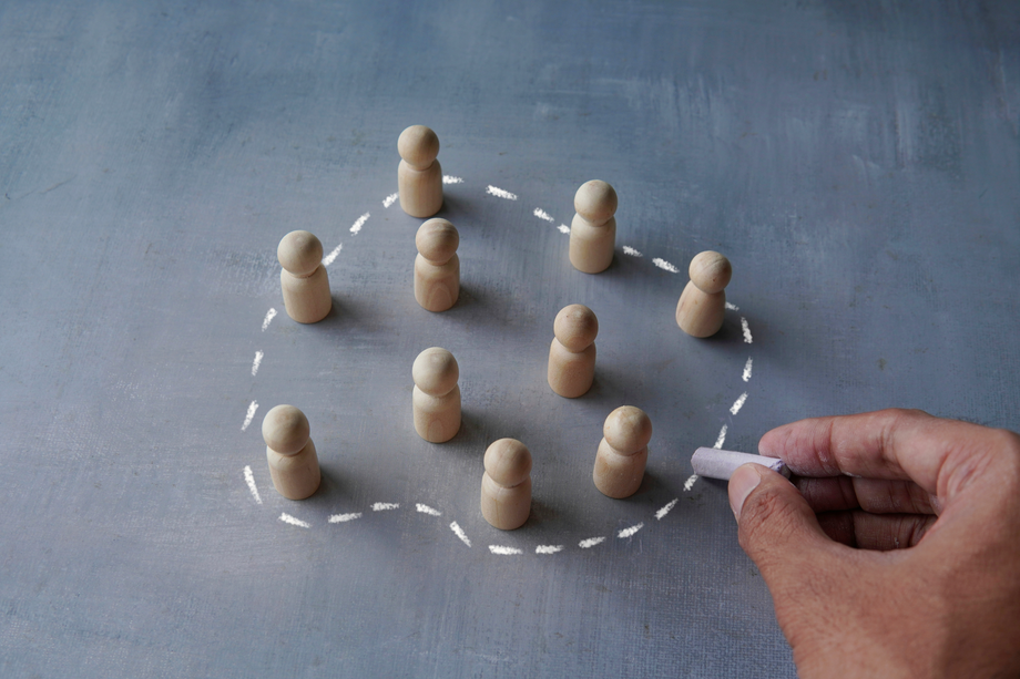 Hand drawing a chalk circle around wooden figurines on a grey surface, illustrating concepts of inclusion, community, or teamwork.