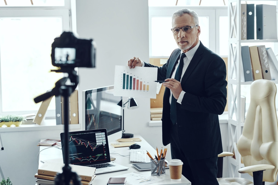 Mature businessman presenting financial data charts to a camera for a business vlog in an office setting