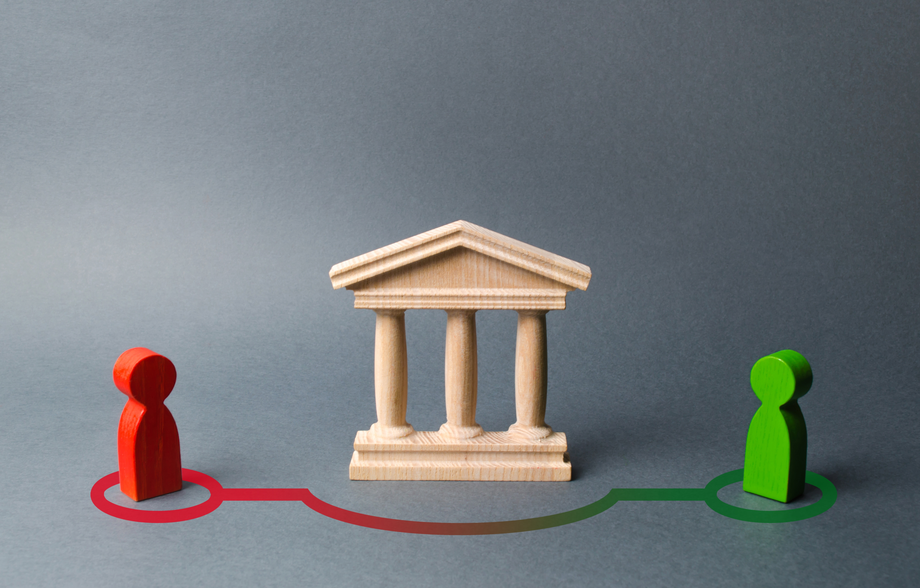 Two wooden figures connected by a red and green pathway to a miniature wooden bank, symbolizing financial planning or investment paths on a gray background.