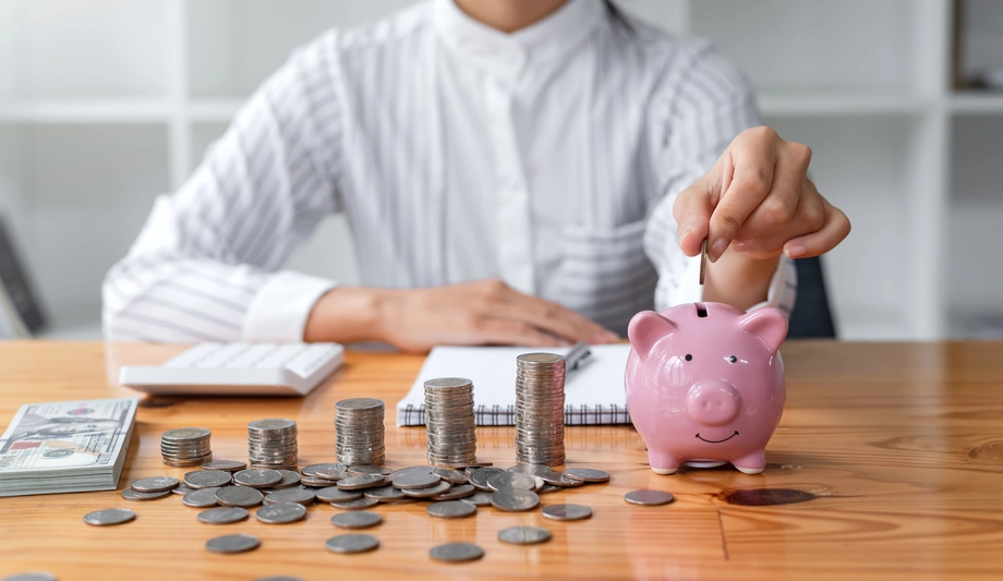 Individual saving money by inserting a coin into a pink piggy bank with stacks of coins and cash on a desk, emphasizing personal financial management.