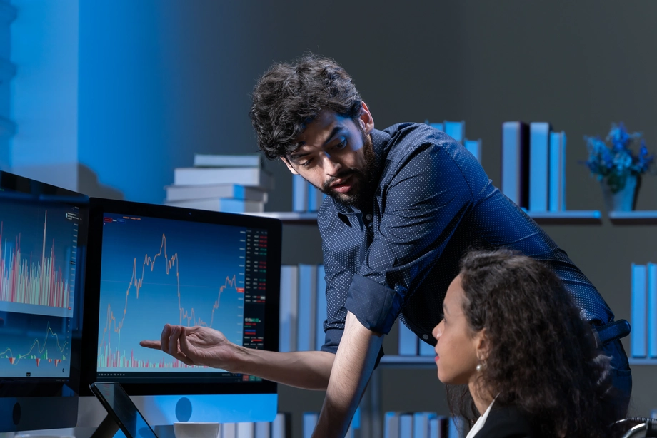 Financial analysts discussing market downturn trends on computer monitors in an office setting.