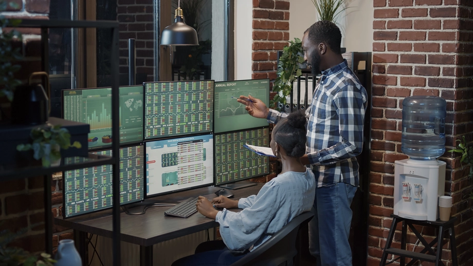 Two individuals analyzing financial data on multiple computer monitors in a modern office with brick walls, representing teamwork in financial analytics.