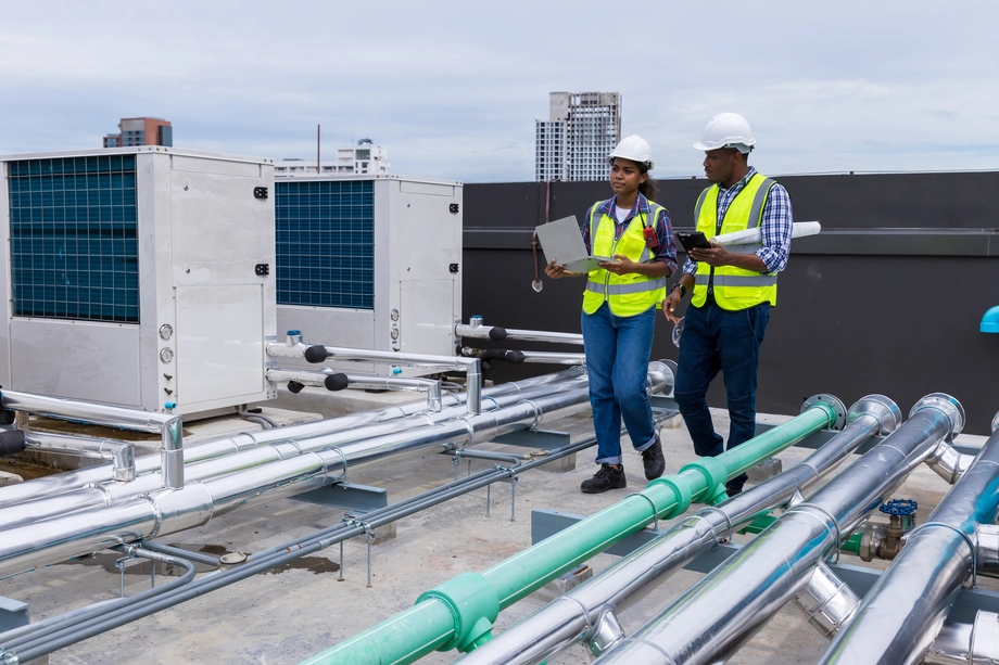 Two engineers with safety helmets and high-visibility vests inspecting HVAC systems on a rooftop, engaged in maintenance and review of industrial equipment.