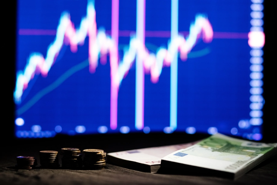 Alt text: "Foreground focus on stacks of coins and euro banknotes with a blurred stock market graph in the background, illustrating financial investment and analysis.