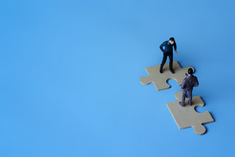 Miniature figures on two connecting puzzle pieces against a blue background, depicting collaboration, partnership, or problem-solving.