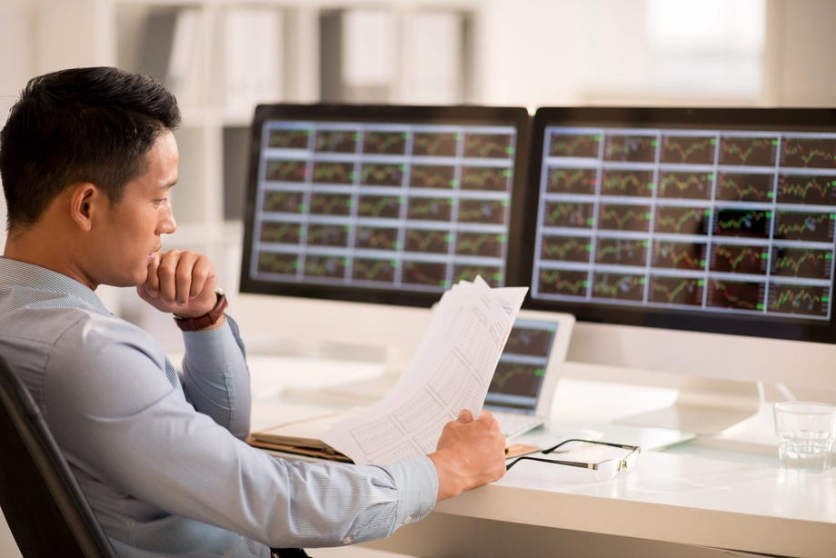 Focused financial analyst reviewing reports with multiple stock market graphs in the background