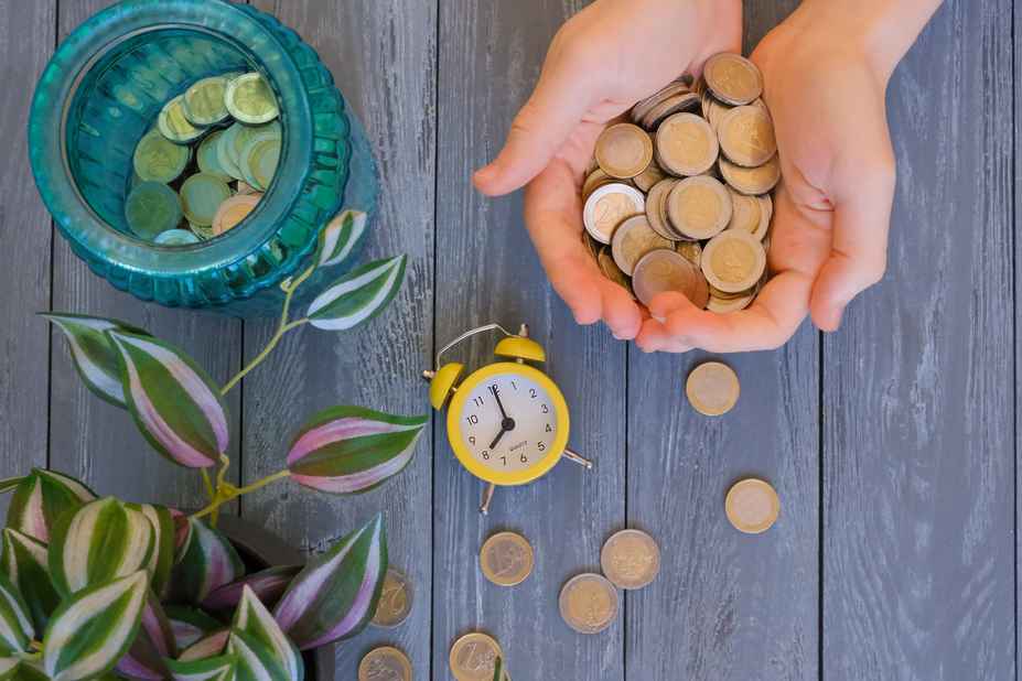 Hands holding coins over a wooden table with a spilled jar of money, a small clock, and a plant, illustrating the concept of liquidity in finance.